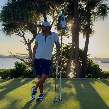 A man wearing a PBGC Course Record Knit Polo in white with dark trim, standing on a golf green at sunset.
