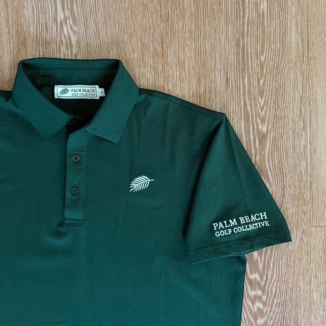 PBGC Pure Strike dark green golf polo shirt with white leaf logo and sleeve branding on a wooden background