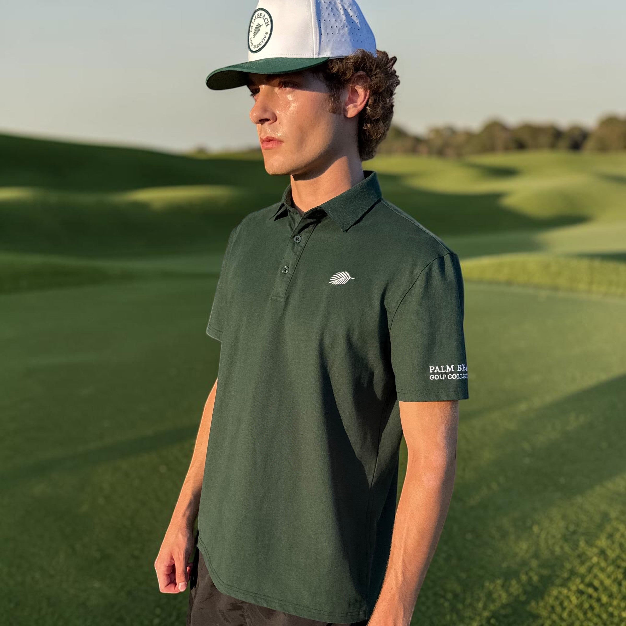 A man wearing a PBGC forest green performance golf polo with a palm leaf logo and white cap on a sunny golf course.