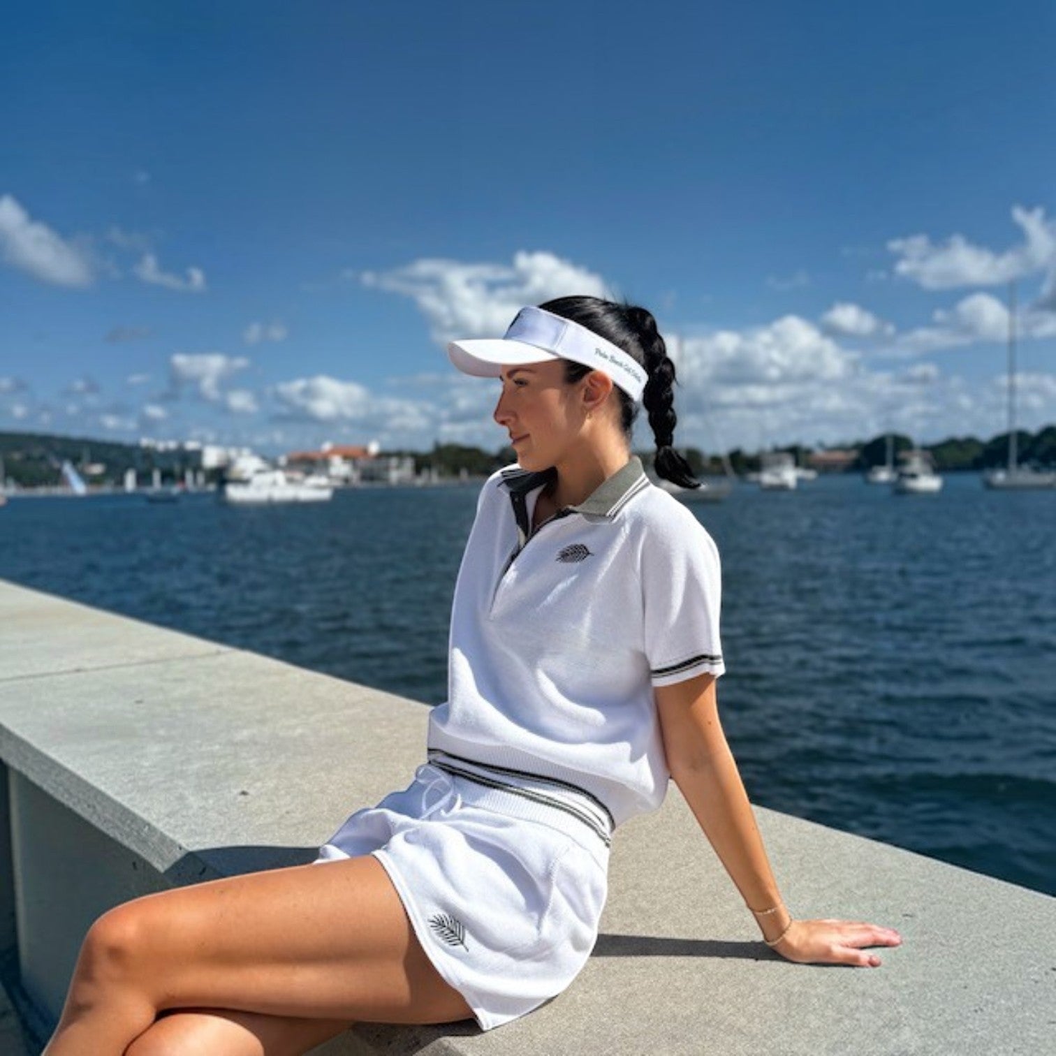 Woman wearing a white PBGC Island Visor and matching white polo set while sitting by the waterfront on a sunny day.