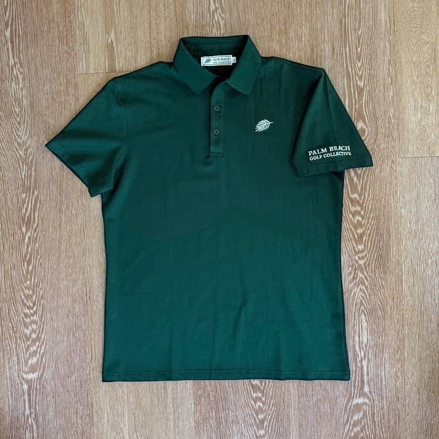 PBGC Pure Strike dark green golf polo shirt with white leaf logo and sleeve embroidery on a wooden background