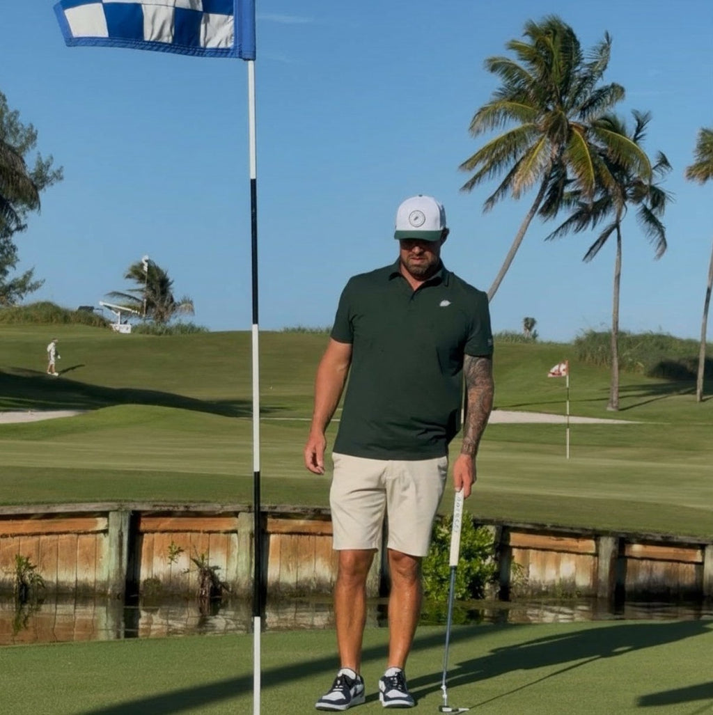 PBGC Pure Strike Polo in dark green worn by a golfer on a sunny putting green with palm trees in the background.
