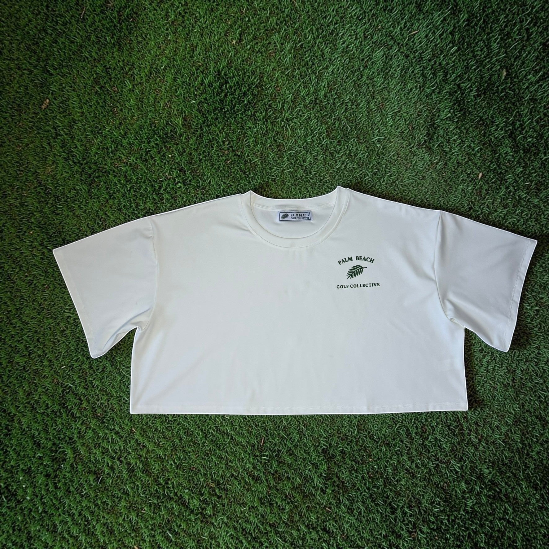 PBGC white crop top with a crew neckline and Palm Beach Golf Collective logo on the chest, laid on green turf.
