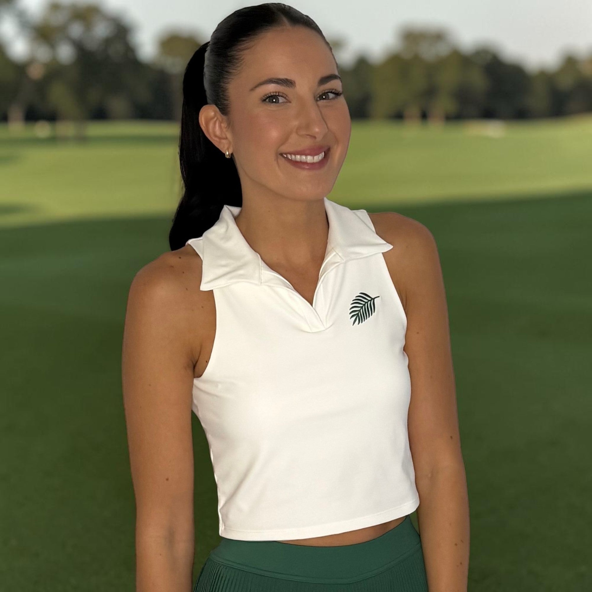 PBGC white sleeveless cropped golf polo with collar and palm leaf embroidery worn by a woman on a golf course