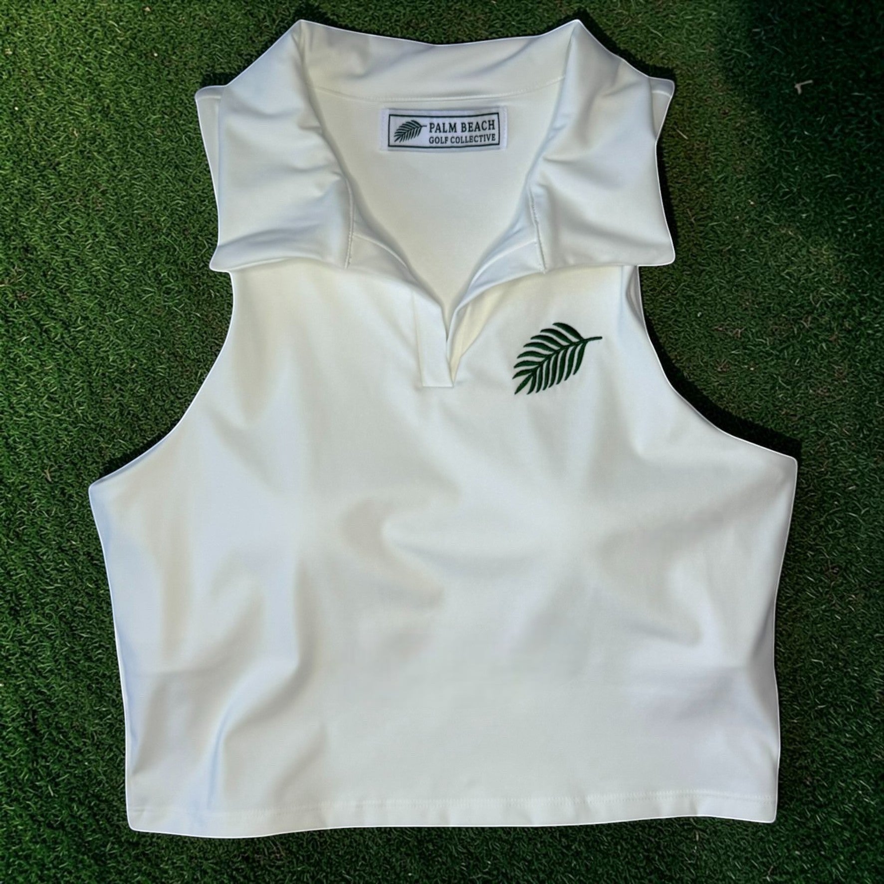 PBGC white sleeveless cropped golf polo with a soft collar, V-neck placket, and green embroidered palm leaf detail.