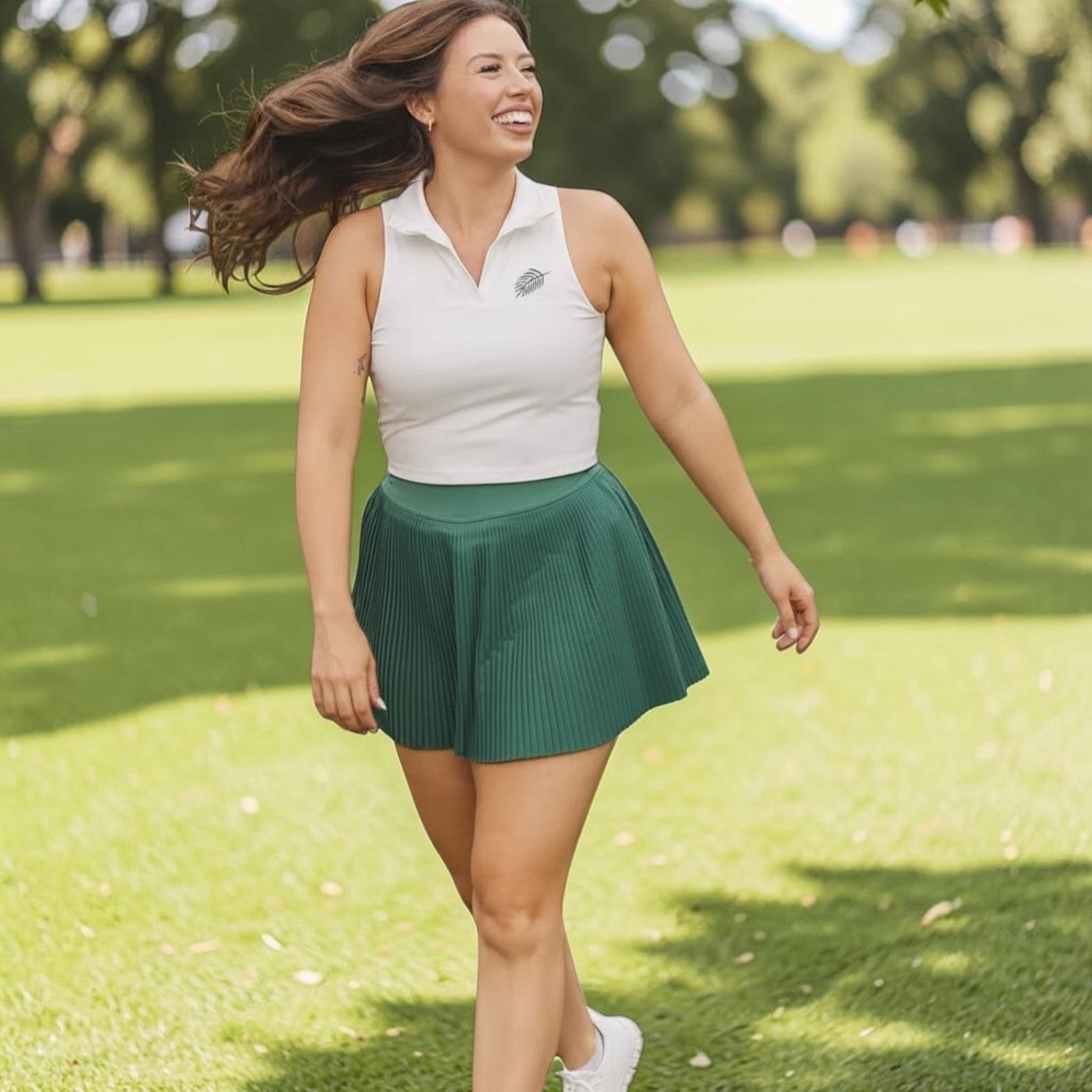 A woman wearing a white PBGC cropped sleeveless polo with a palm leaf detail and a green pleated golf skirt.