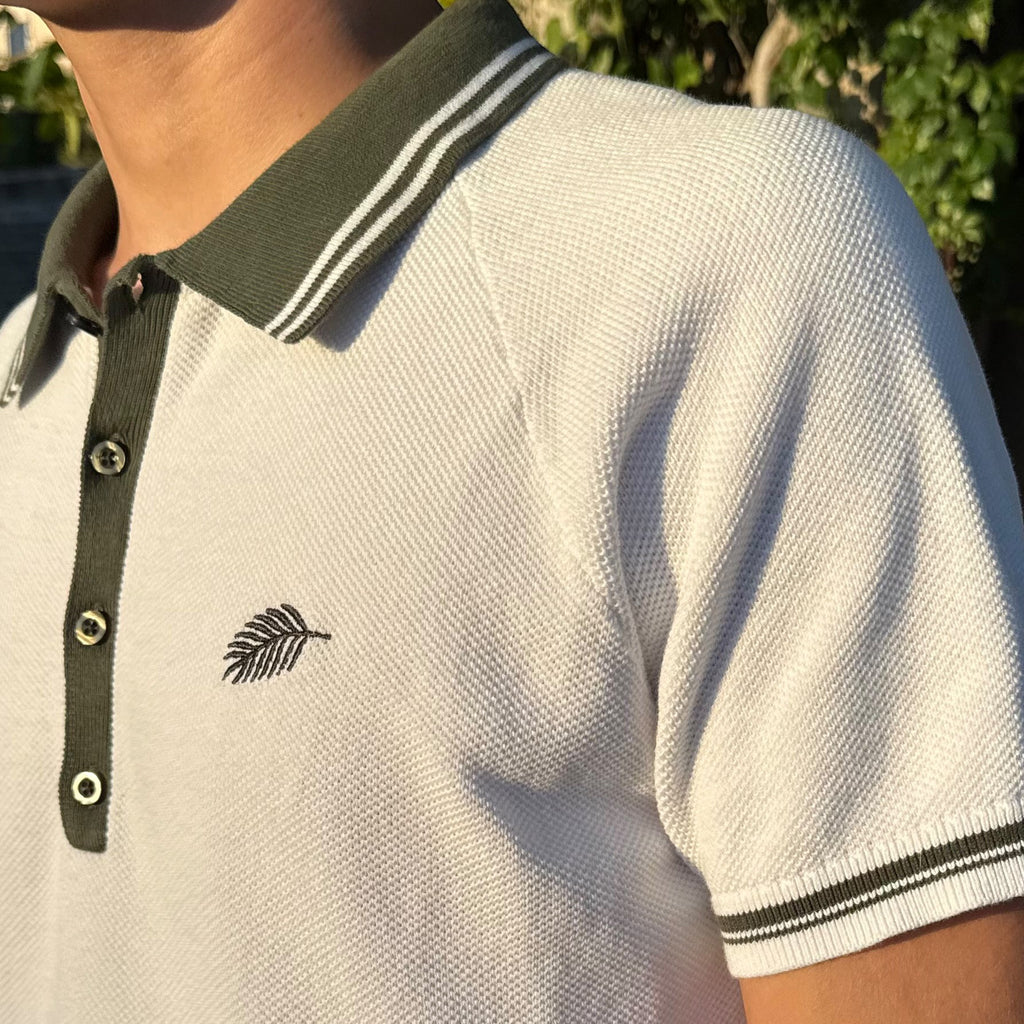 PBGC white cotton knit polo shirt with olive green striped collar, raglan sleeves, and palm leaf chest embroidery.