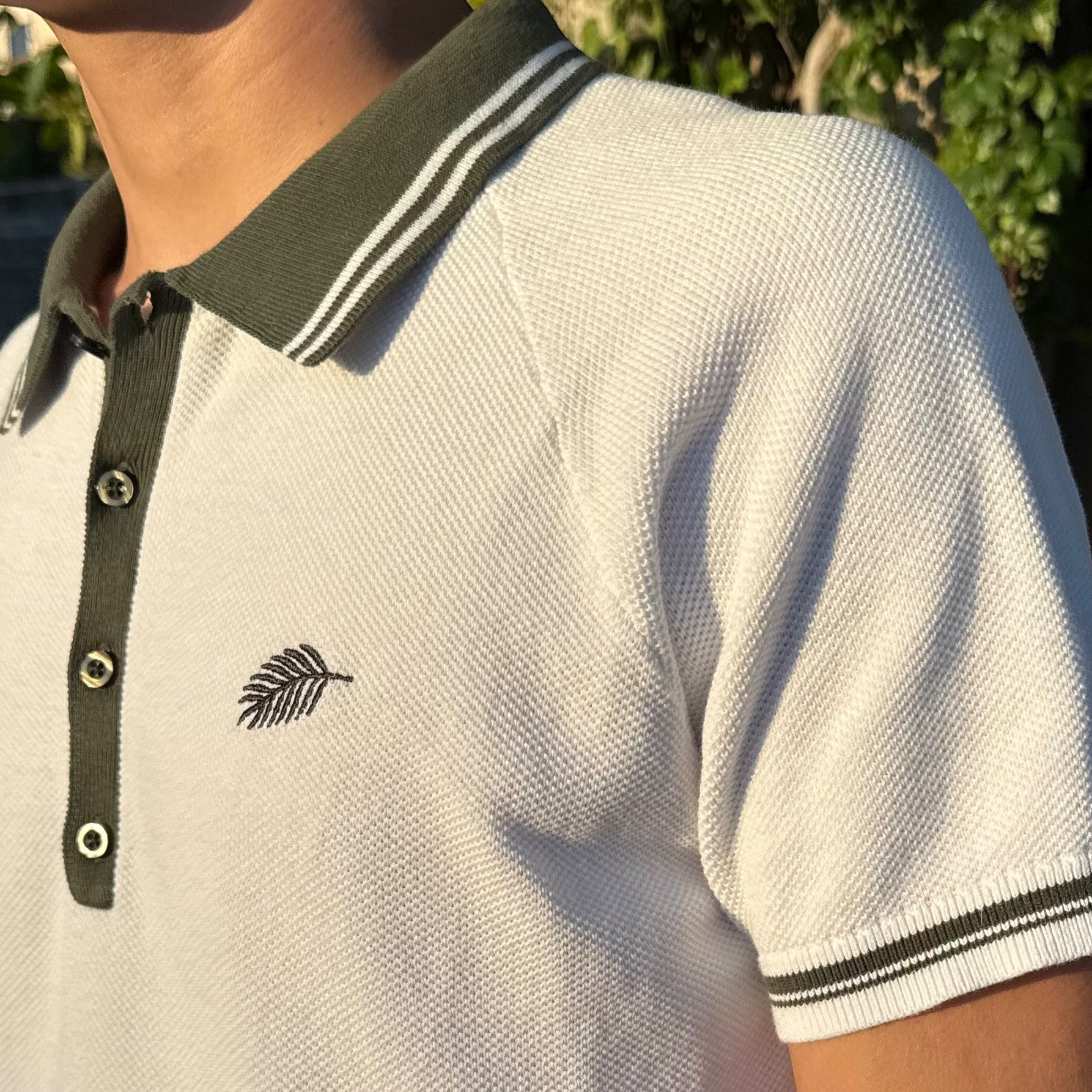 PBGC white cotton knit polo shirt with olive green striped collar, raglan sleeves, and palm leaf chest embroidery.