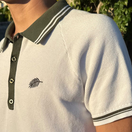 PBGC white cotton knit polo shirt with olive green striped collar, raglan sleeves, and palm leaf chest embroidery.