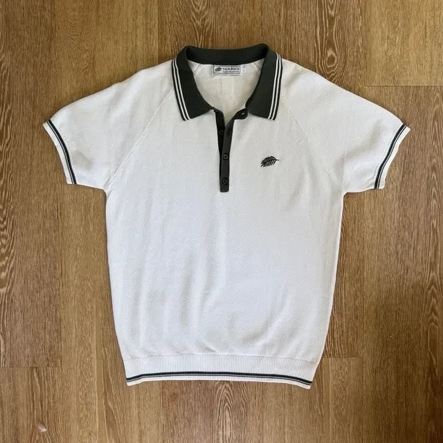PBGC white knit polo shirt with olive green collar, striped trim, and palm leaf embroidery on the chest.