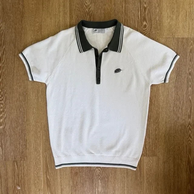 PBGC white cotton knit polo with olive green collar, striped trim, and palm leaf embroidery on the chest.