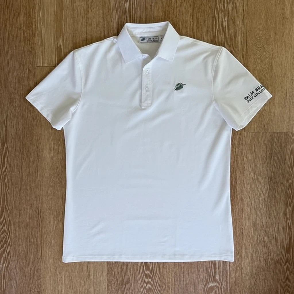 PBGC One Under Par white performance golf polo with a green palm leaf logo and sleeve branding on a wood background.