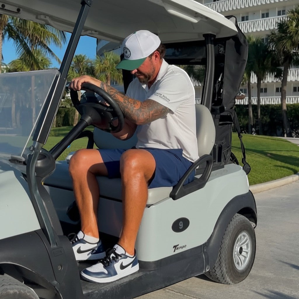 PBGC golfer in a white performance polo and navy shorts driving a golf cart on a sunny day.
