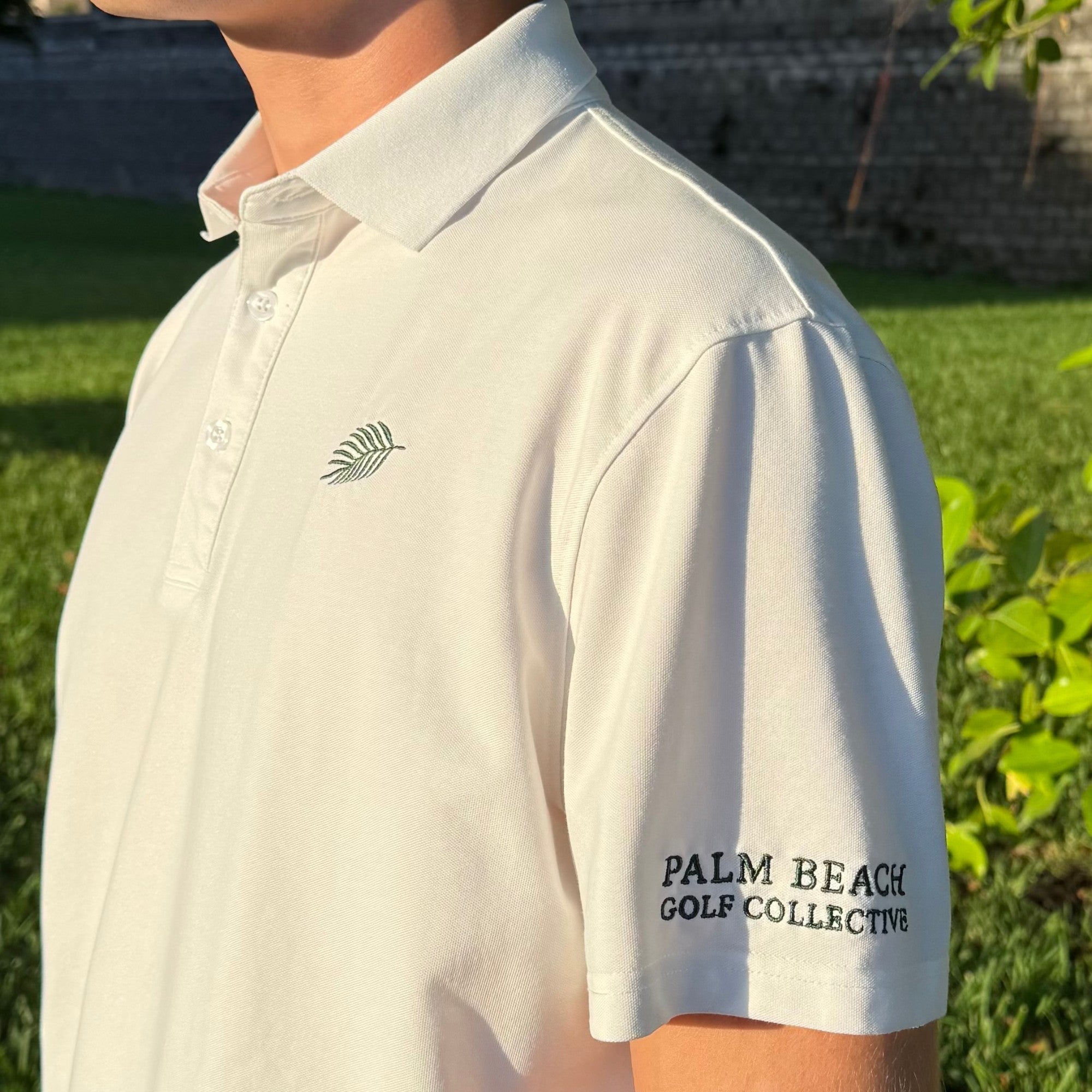 PBGC white performance golf polo with green palm leaf chest logo and sleeve branding worn outdoors