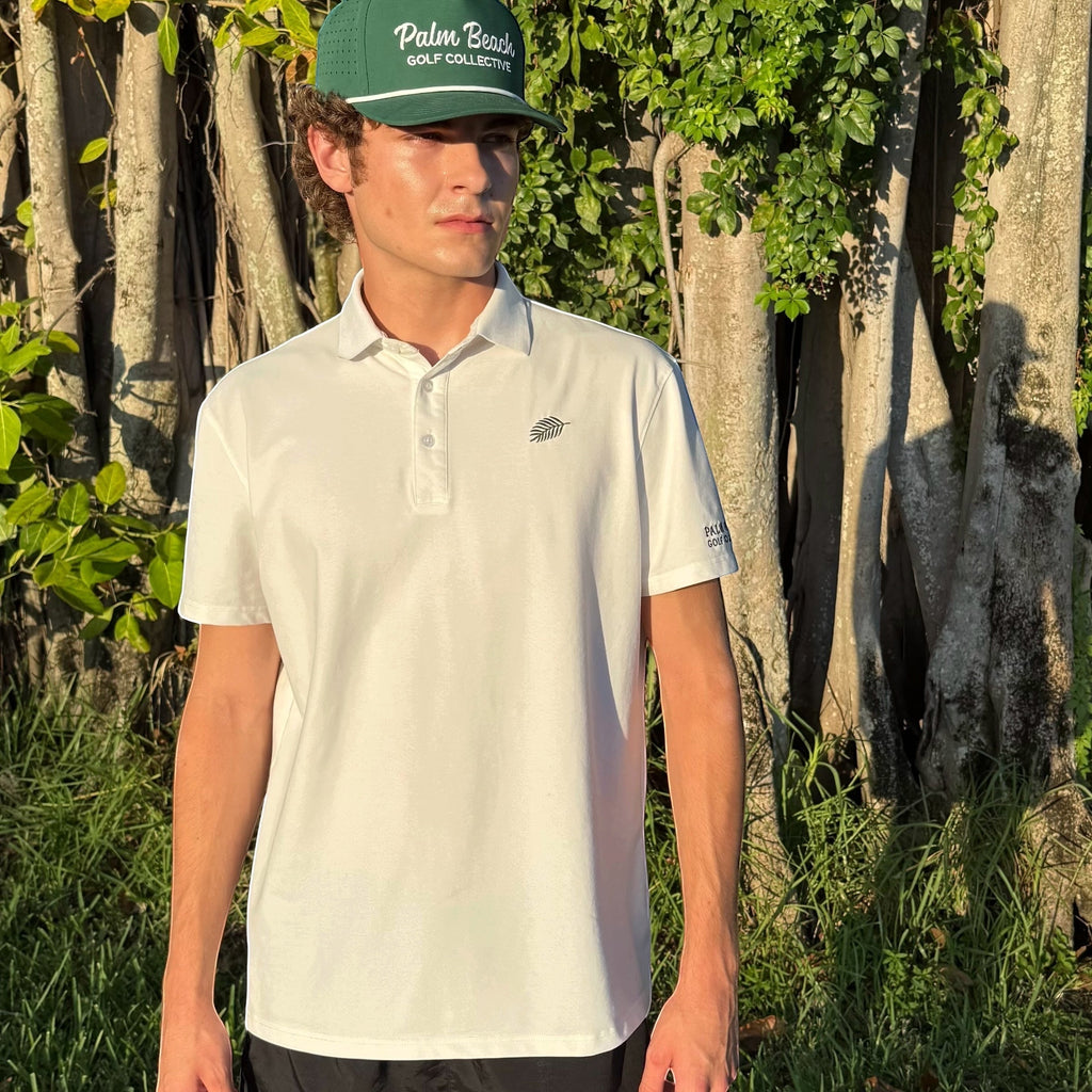 A man wearing a PBGC white performance polo shirt and a green Palm Beach Golf Collective cap outdoors.