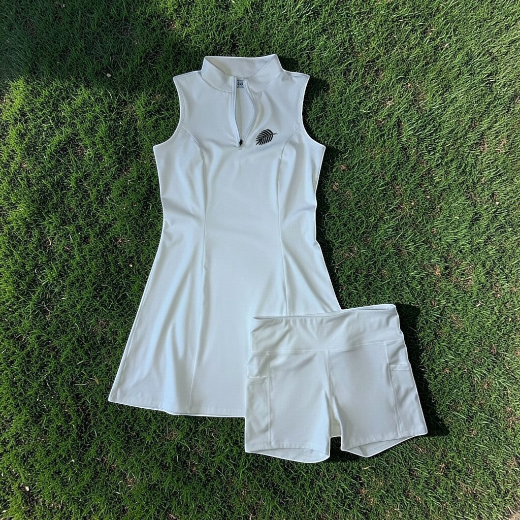 PBGC white sleeveless performance golf dress with mock neck, quarter-zip, and matching undershorts on green grass.