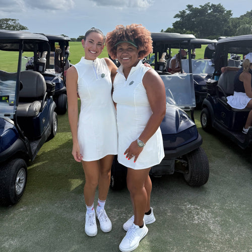 Two women wearing white PBGC sleeveless performance golf dresses with mock-neck collars on a sunny golf course.